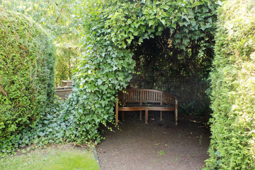 Holzbank unter einem grünen Laubbogen im Garten, umgeben von Hecken