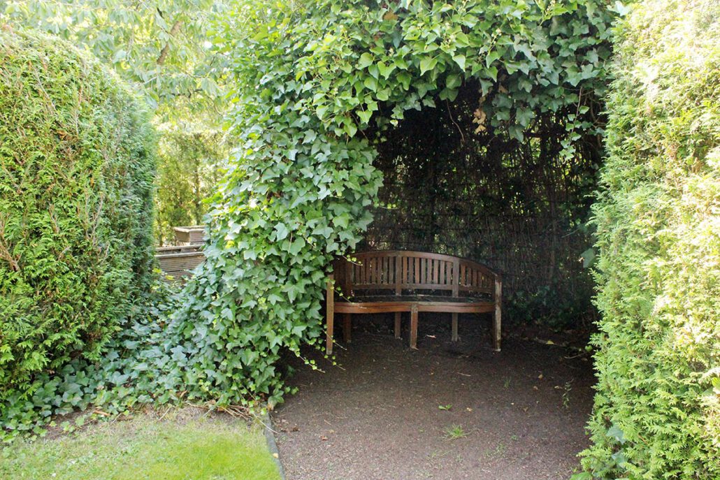 Holzbank unter einem grünen Laubbogen im Garten, umgeben von Hecken