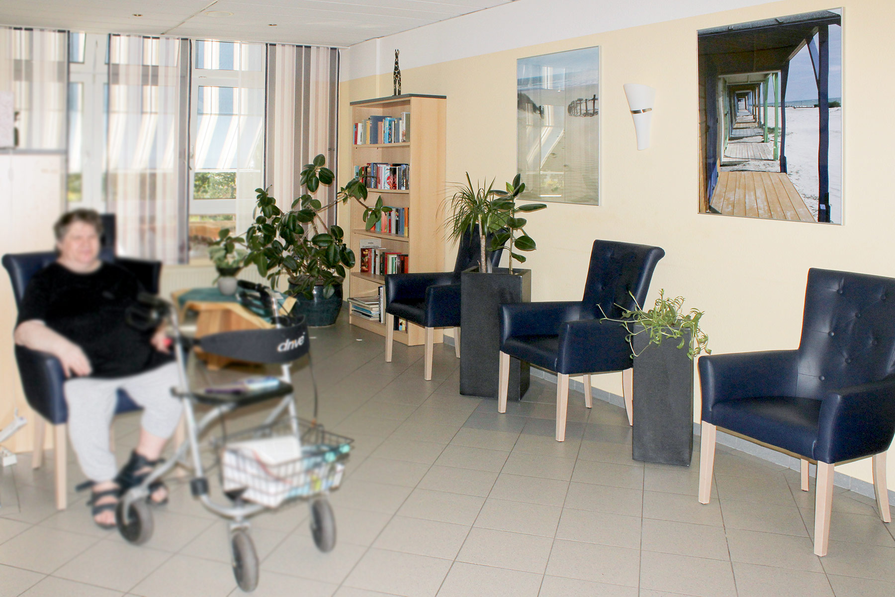 Bewohnerin sitzt mit Rollator in einem hellen Gemeinschaftsraum mit blauen Sesseln, Pflanzen und Bücherregal